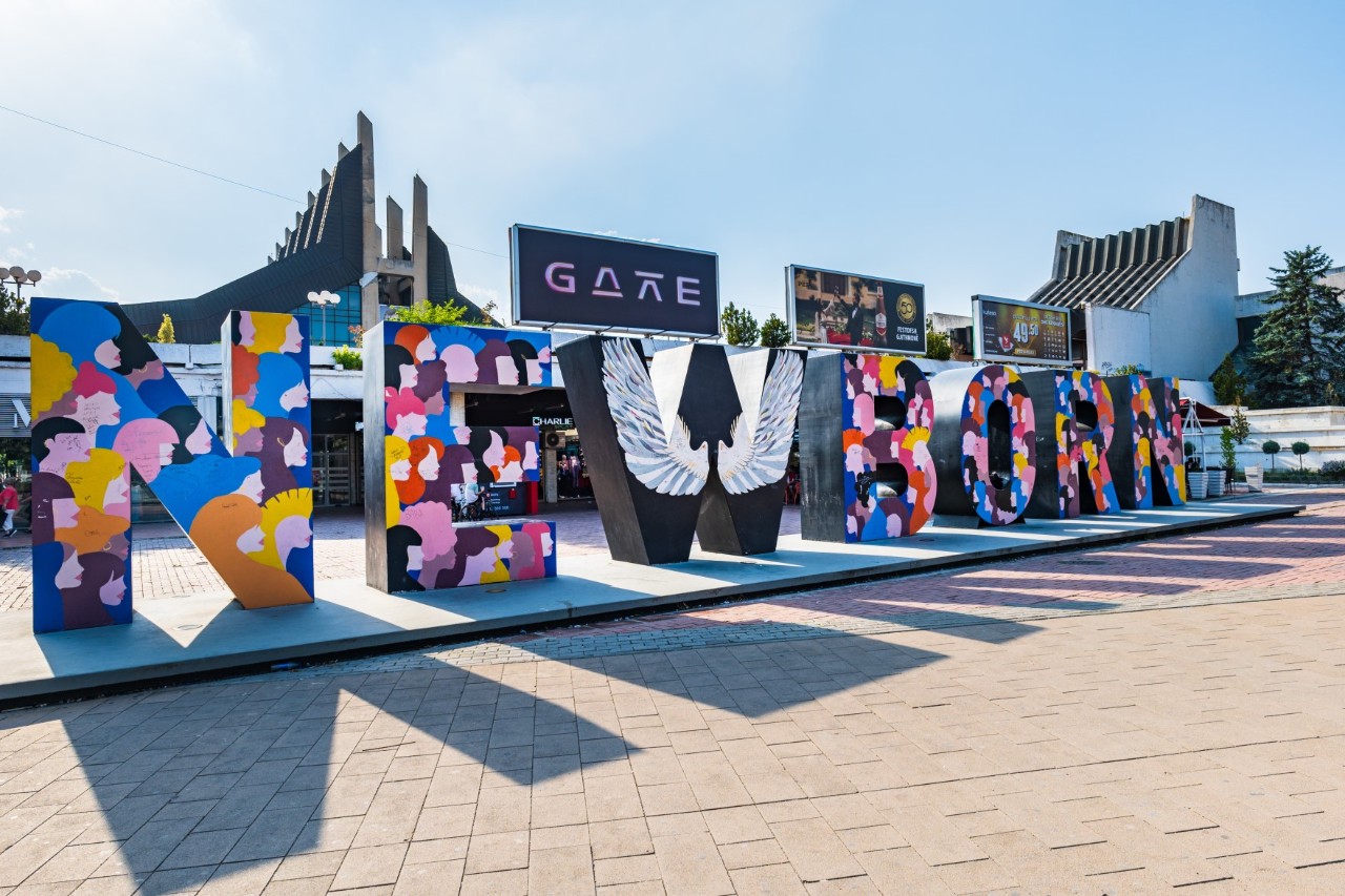 Large paved square with a sculpture made up of individual letters spelling out the word NEWBORN. The letters are painted in bright colours. Behind the sculpture stands a futuristic building.