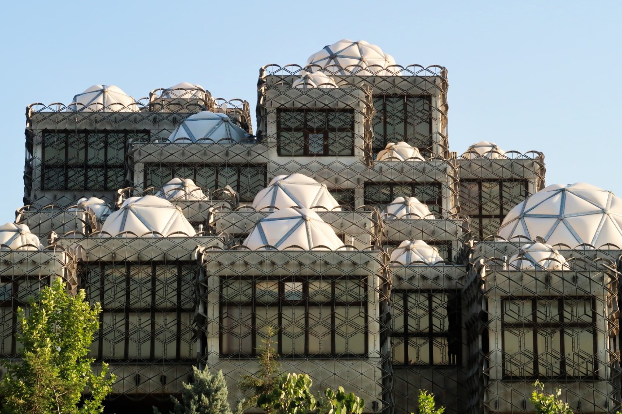 A large concrete building made up of many individual blocks, each covered by a white dome. The parts of the building are enclosed by metal grids with geometric shapes. The tall windows have black metal frames.