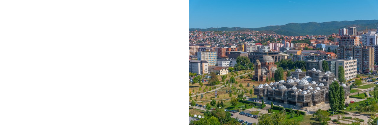 View of the city from above. In the foreground, green lawns, trees, and several paved areas can be seen. Closest to the viewer is a large, jagged, grey structure with numerous dome-shaped towers, behind which rise many multi-storey houses. A chain of hills can be seen in the background.
