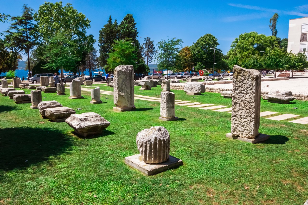 The remains of columns and buildings are arranged geometrically on a green meadow. A path laid out with stone slabs leads through the columns. Various trees can be seen in the background.