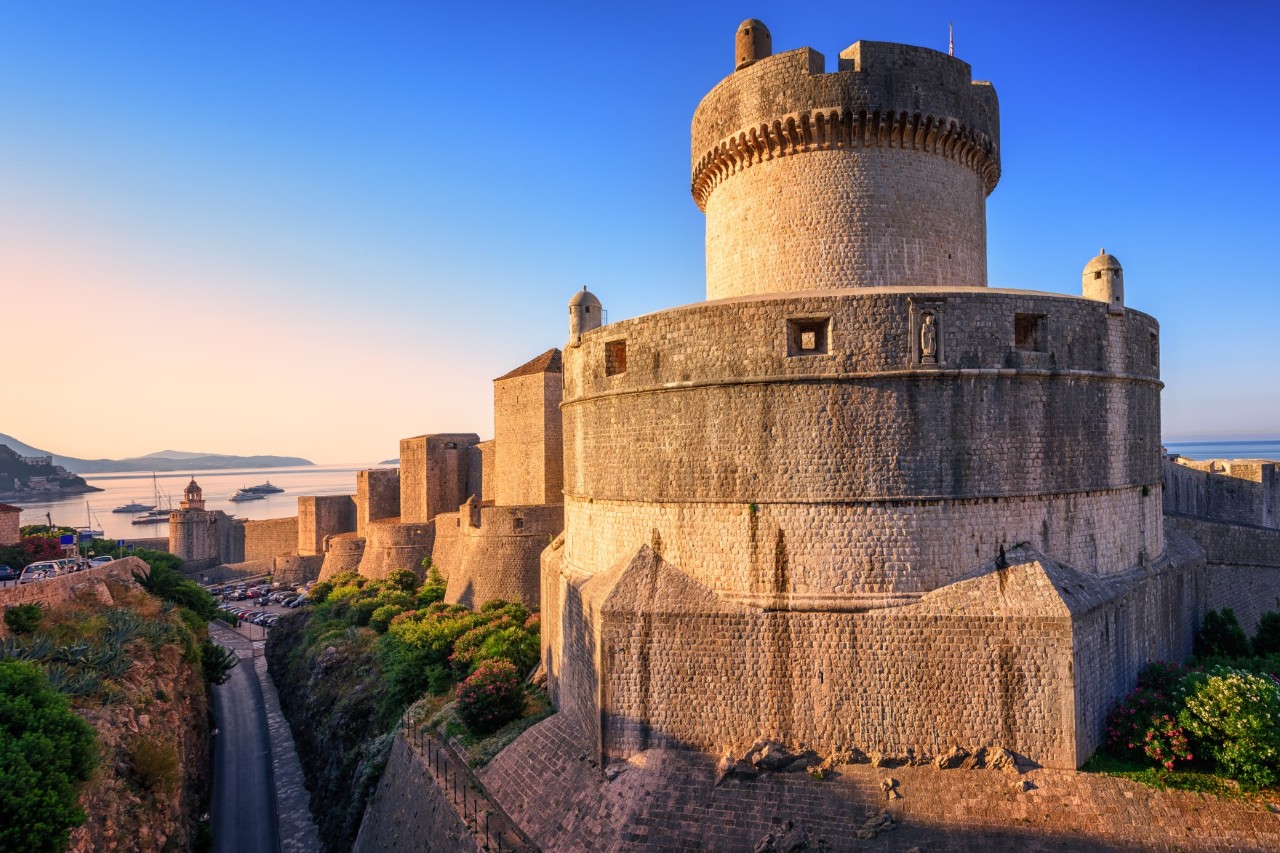 A huge, circular fortress with a round castle tower in the centre in the evening light. The fortress is part of a larger fortification that can be seen in the background. Trees and bushes can be seen below the fortress walls. The sea and parts of the coast are visible in the background.
