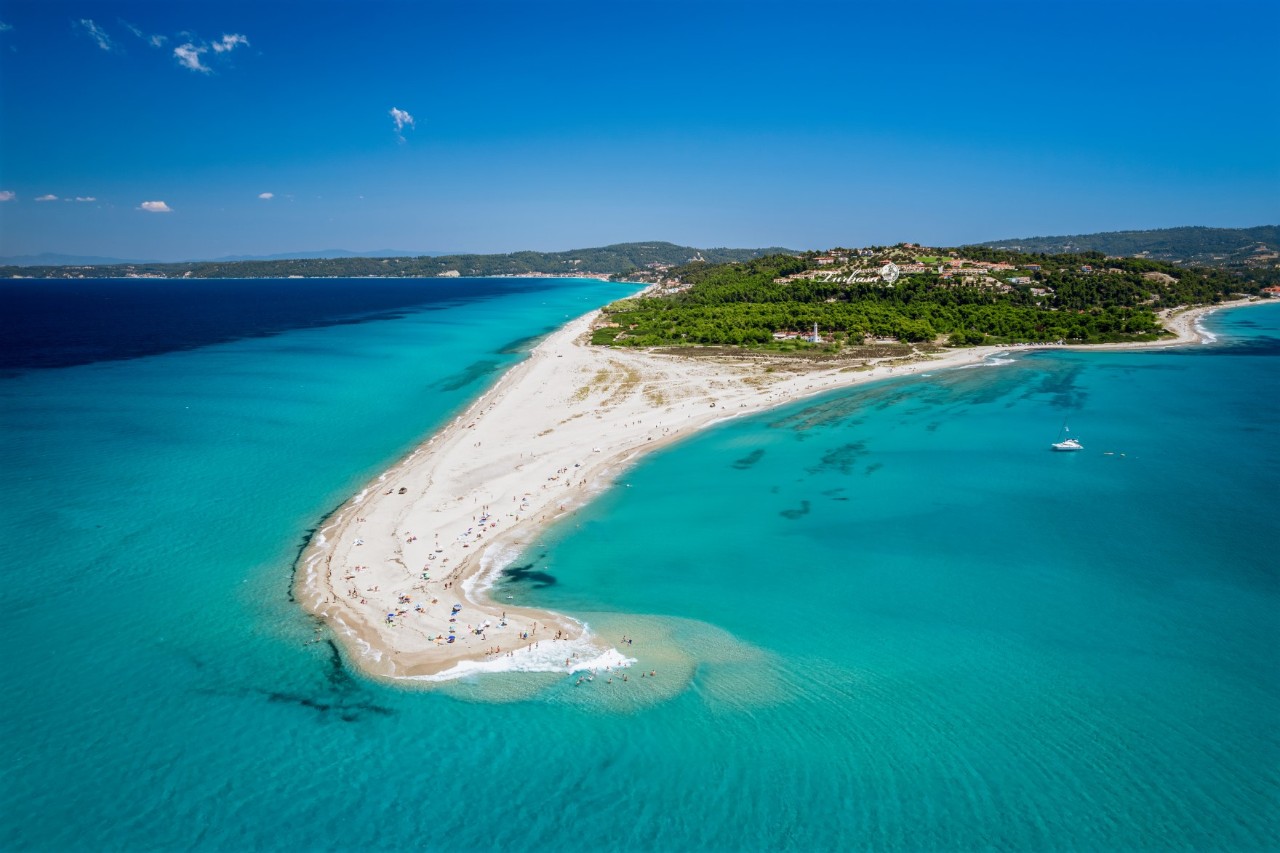An elongated headland completely covered with sand, jutting out into the shallow, turquoise sea. A green, forested landscape with rolling hills can be seen in the background.