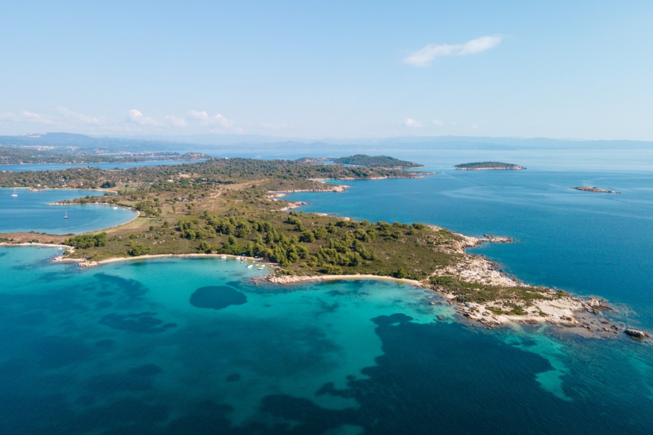 An aerial view of a coastal landscape with several small bays and clear turquoise water. The coast is covered by green vegetation.