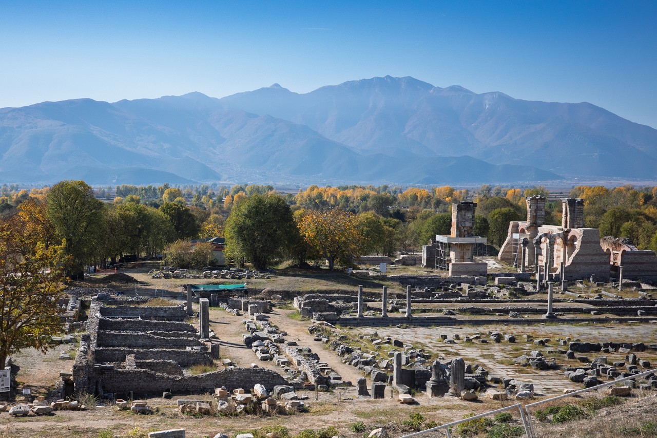 Extensive archaeological site with ancient columns, stone paths, and remains of historic buildings. In the background, green countryside and mountains stretch out under a bright sky.