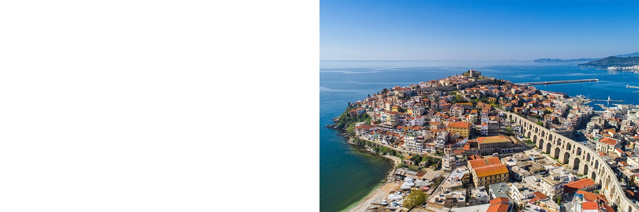 Aerial view of a coastal town with densely packed houses with red roofs on a peninsula. On the right side of the image, a large, historic aqueduct runs through the town. In the background, the sea, a harbour and rolling hills can be seen under a clear blue sky.