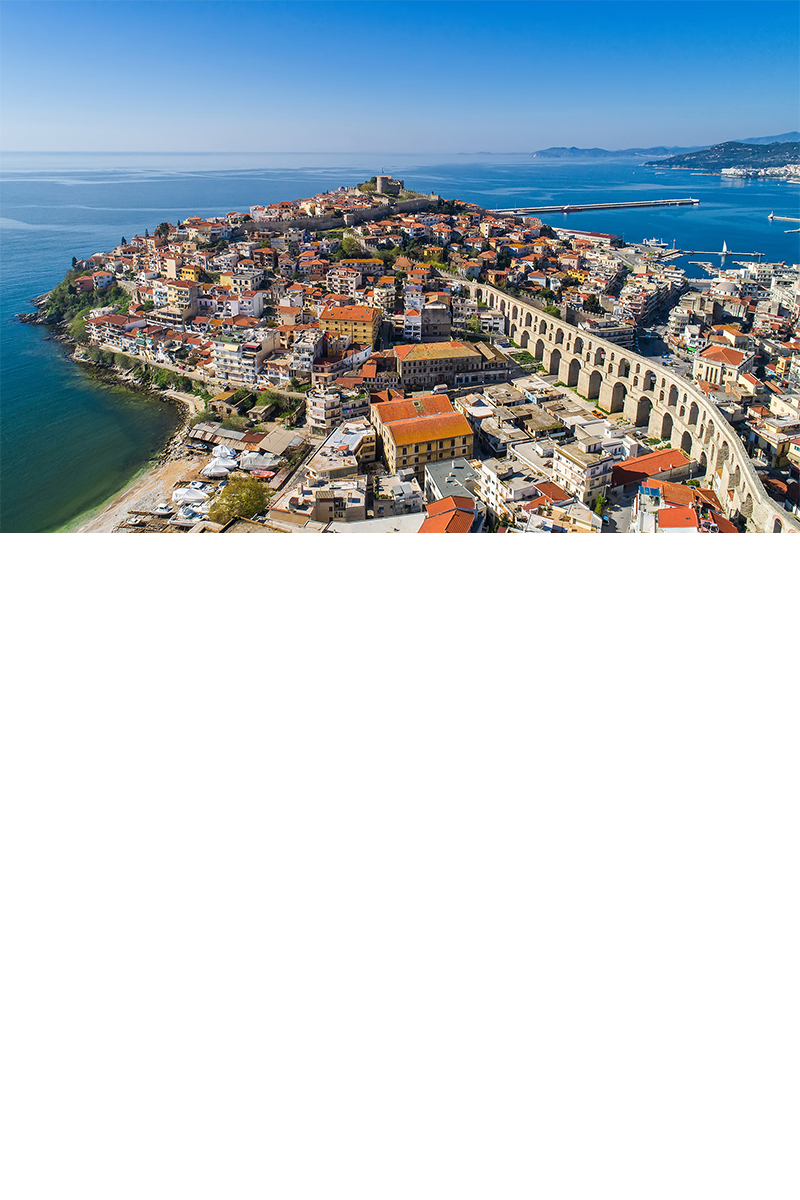 Aerial view of a coastal town with densely packed houses with red roofs on a peninsula. On the right side of the image, a large, historic aqueduct runs through the town. In the background, the sea, a harbour and rolling hills can be seen under a clear blue sky.