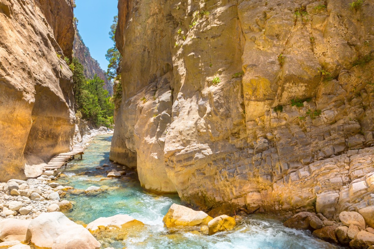 A rushing brook with turquoise-blue water and larger stones in it. To the left and right of the water, huge, steep, ochre-coloured rock walls tower up. 
