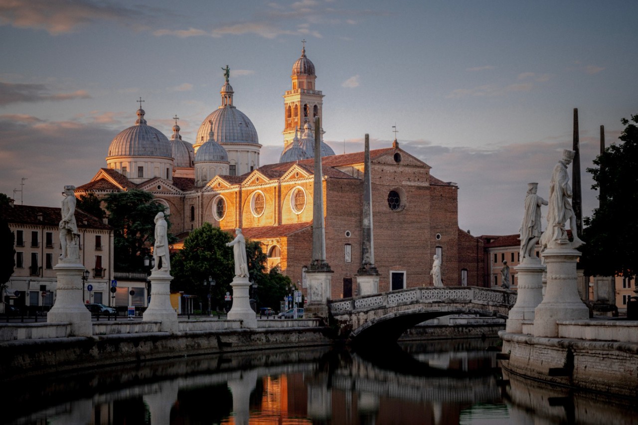 In the foreground, you can see a canal at dusk. To the right and left of the canal are white statues. In the background, there is a church with several domes, illuminated by the sun. 
