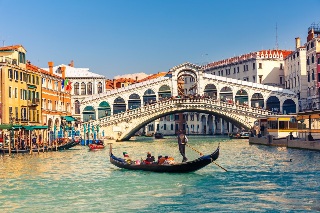 A gondola is travelling through a canal. In the background, there is a large, white arched bridge. On the left side, there is a yellow house façade, and on the right side, a white one. The sky is cloudless, and the sun is shining on the water. 
