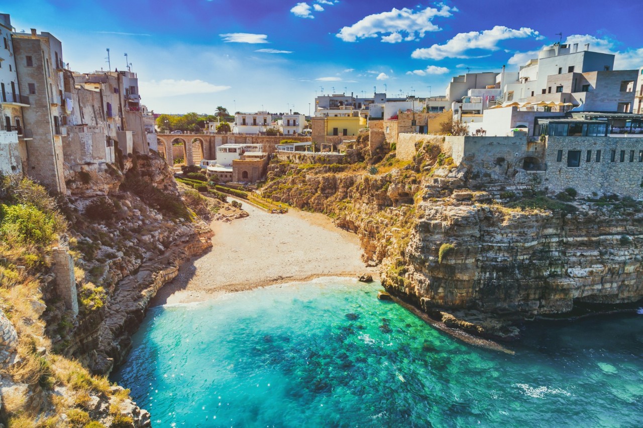 A bay with a sandy beach and turquoise blue water, surrounded by cliffs topped with Mediterranean buildings. 