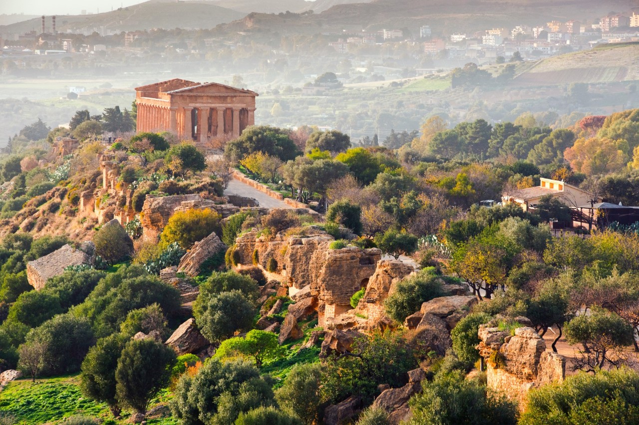 Greek temple with columns and tympanum in a green, hilly valley, buildings in the background 