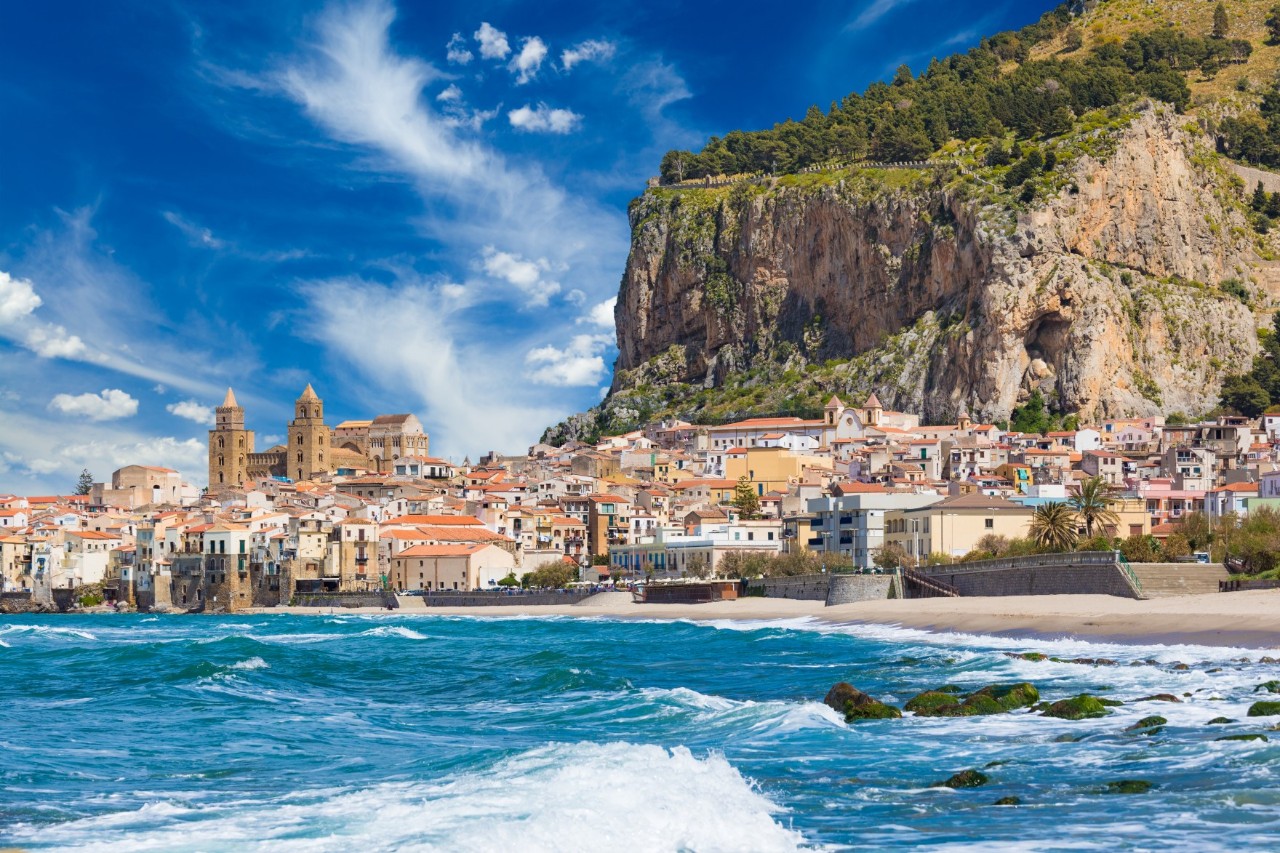 Lots buildings and a cathedral next to the sea.. Behind the town on the right is a towering, green rock massif 
