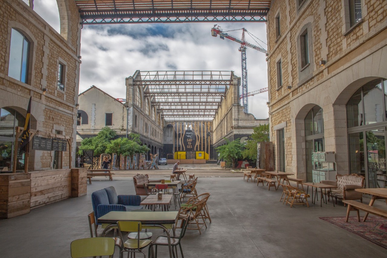 A large, open inner courtyard with buildings with large arched windows to the left and right at the front of the picture. In front of the building on the right are tables, chairs and wooden benches. In the middle of the courtyard are café tables made up of a mix of colourful furniture, from wicker chairs to sofas and tables. At the back of the picture there is a second empty courtyard under a large roof structure. There is a crane behind it.