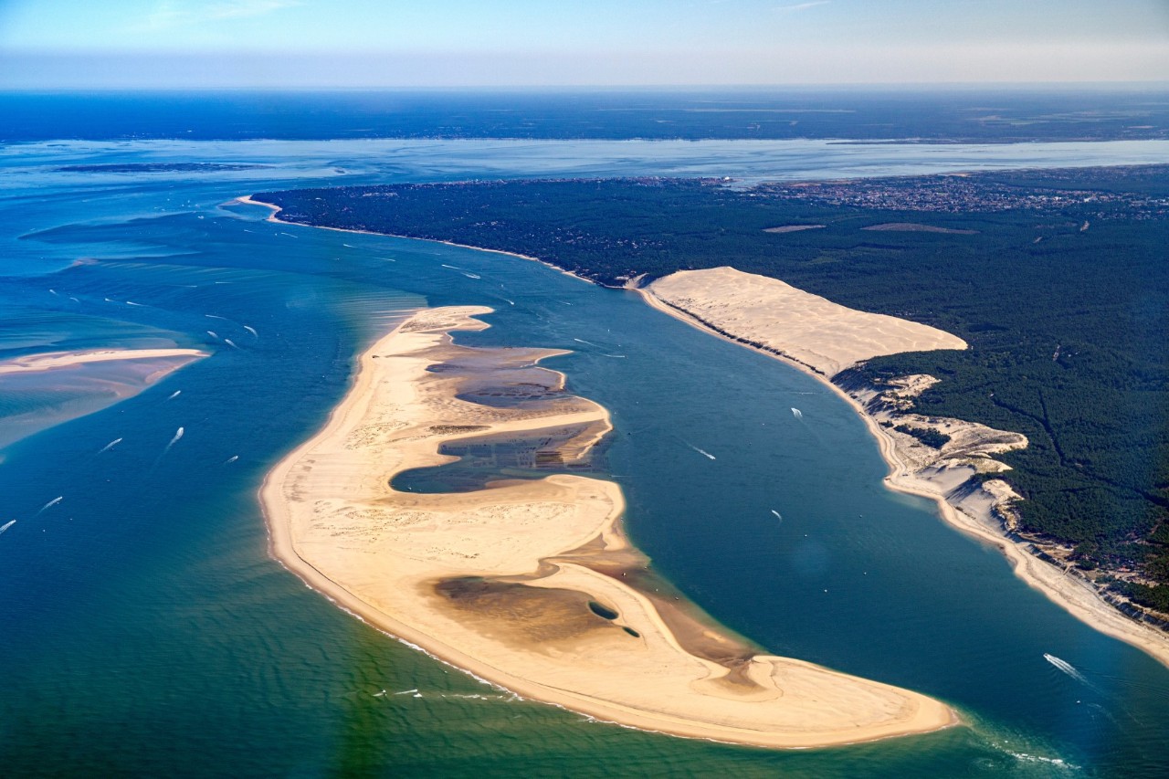 A headland separated by the sea from the mainland on the right. The headland seems to consist completely of sand with large sandy beaches surrounded by dense woodland on the mainland. The sea on both sides of the headland is dark green to dark blue.