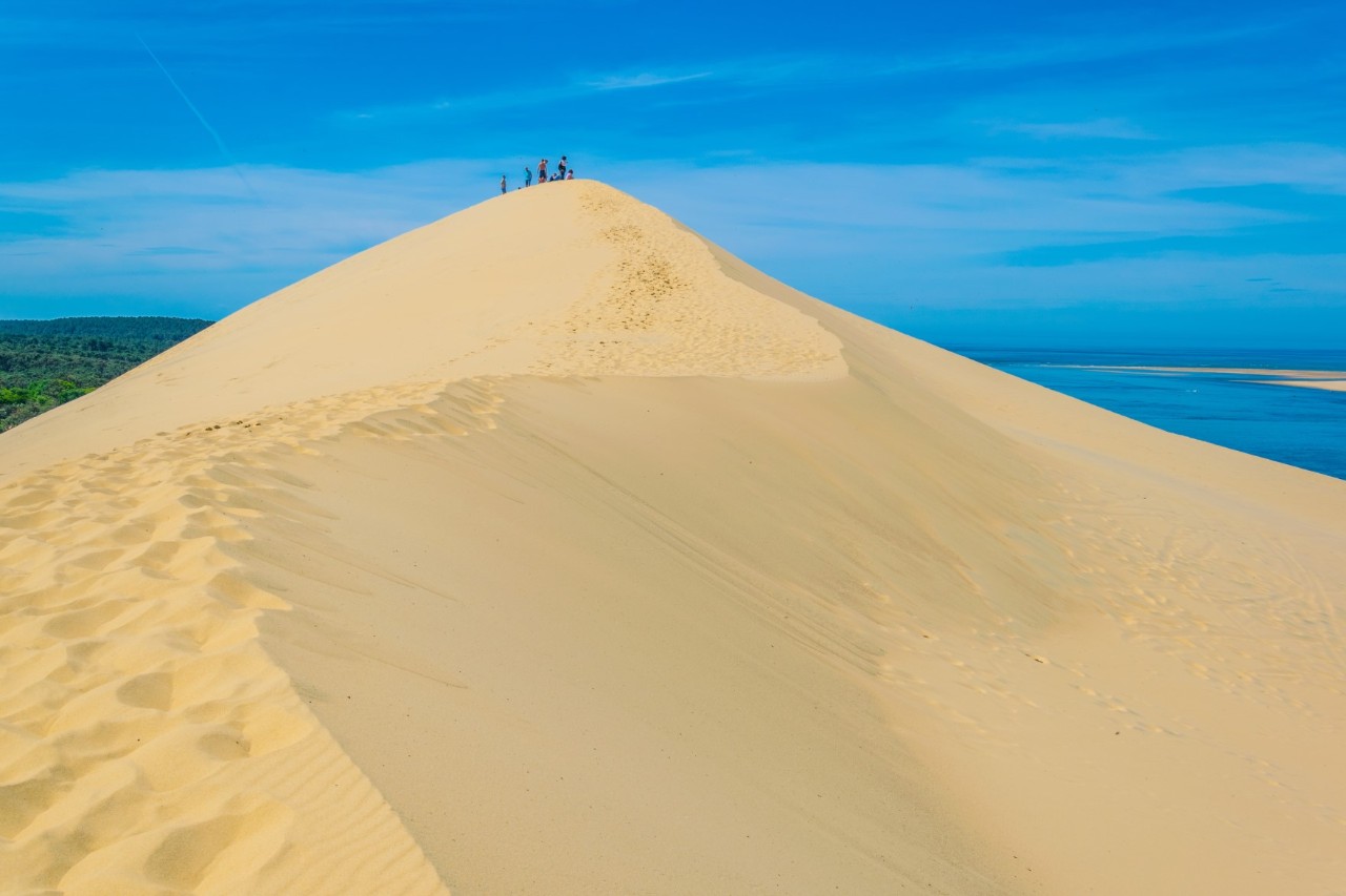 A large sand dune that points like a mountain peak in the centre of the picture. A few people can be seen at the top, with the sea on the right and forest on the left on the horizon. The sky over the dune is blue.