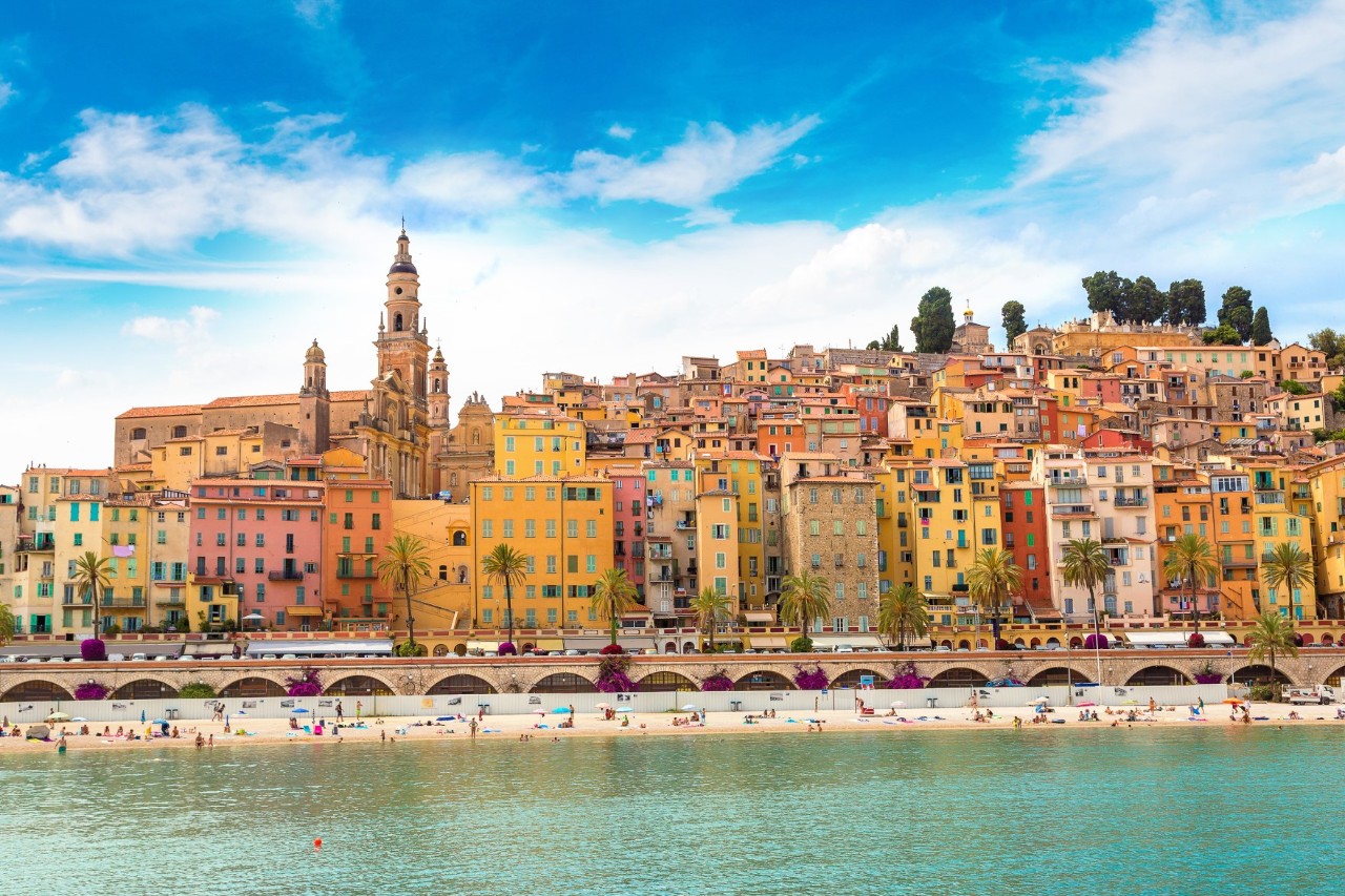 The colourful houses and church tower of Menton, with the beach and water in front of it 