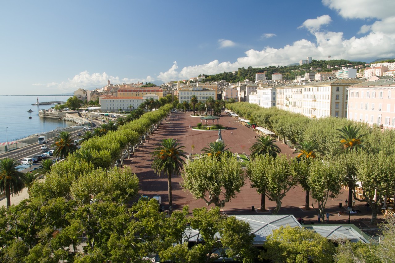 Large, red square surrounded by palm trees and white buildings. On the left is the sea and a promenade frequented by cars.