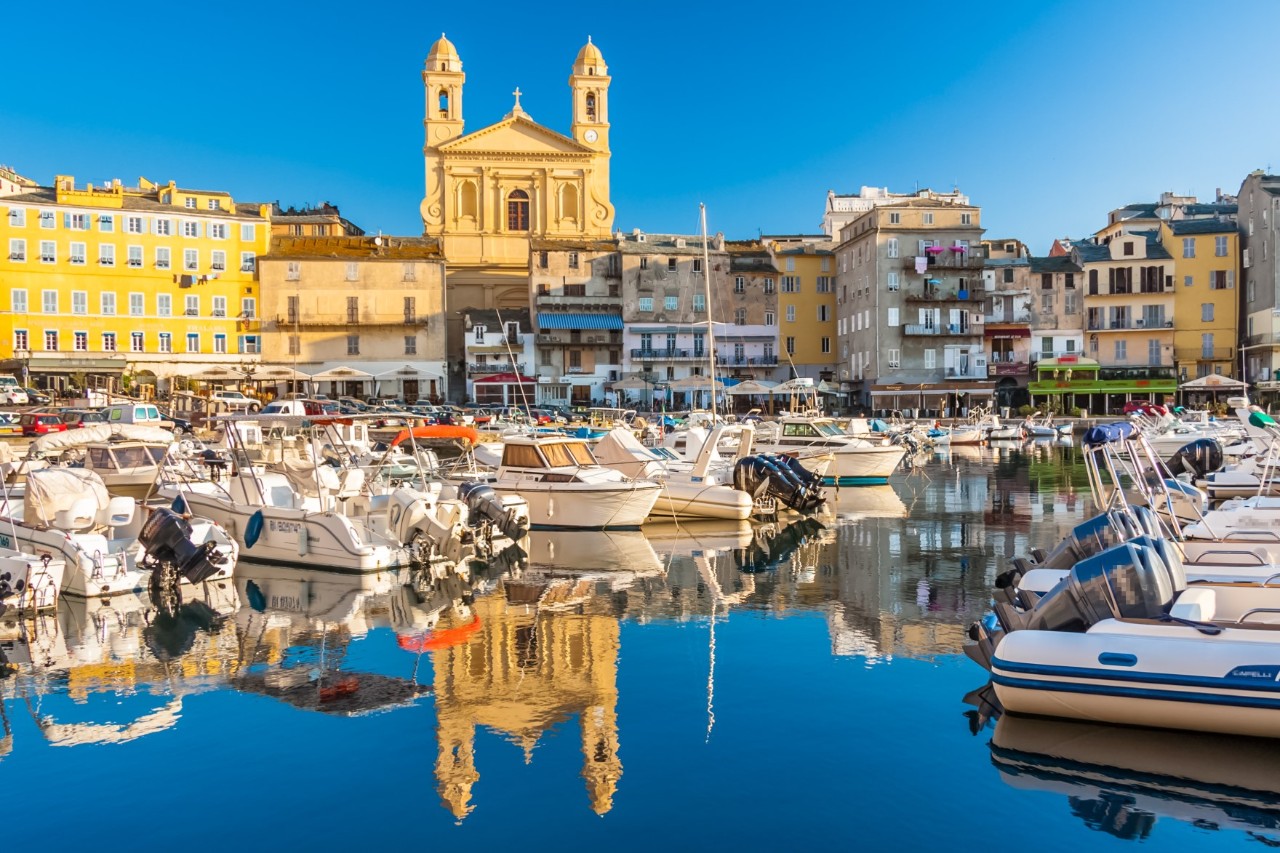 Harbour basin with many small boats to a striking, yellow church with two towers. The church is surrounded by several buildings, which are reflected in the water of the harbour together with the church.