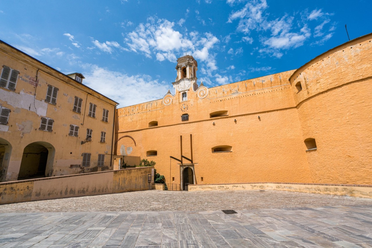 An orange-coloured fortress wall around a deserted square. Next to it is a three-storey building with an archway.