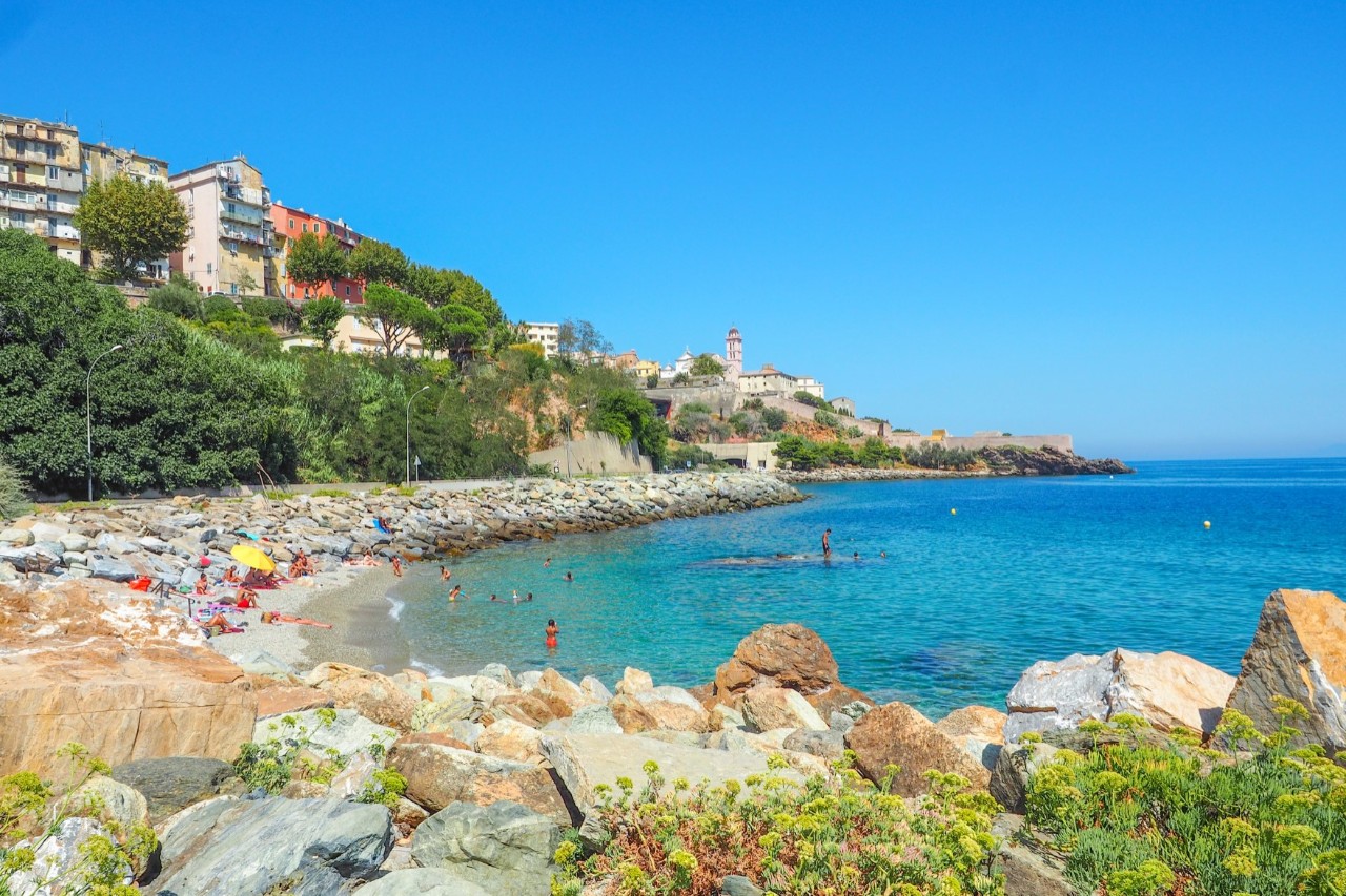 A small bay with few people in the turquoise water. The beach is surrounded by stones in the foreground. Behind the beach is a hill with trees and buildings upon it.