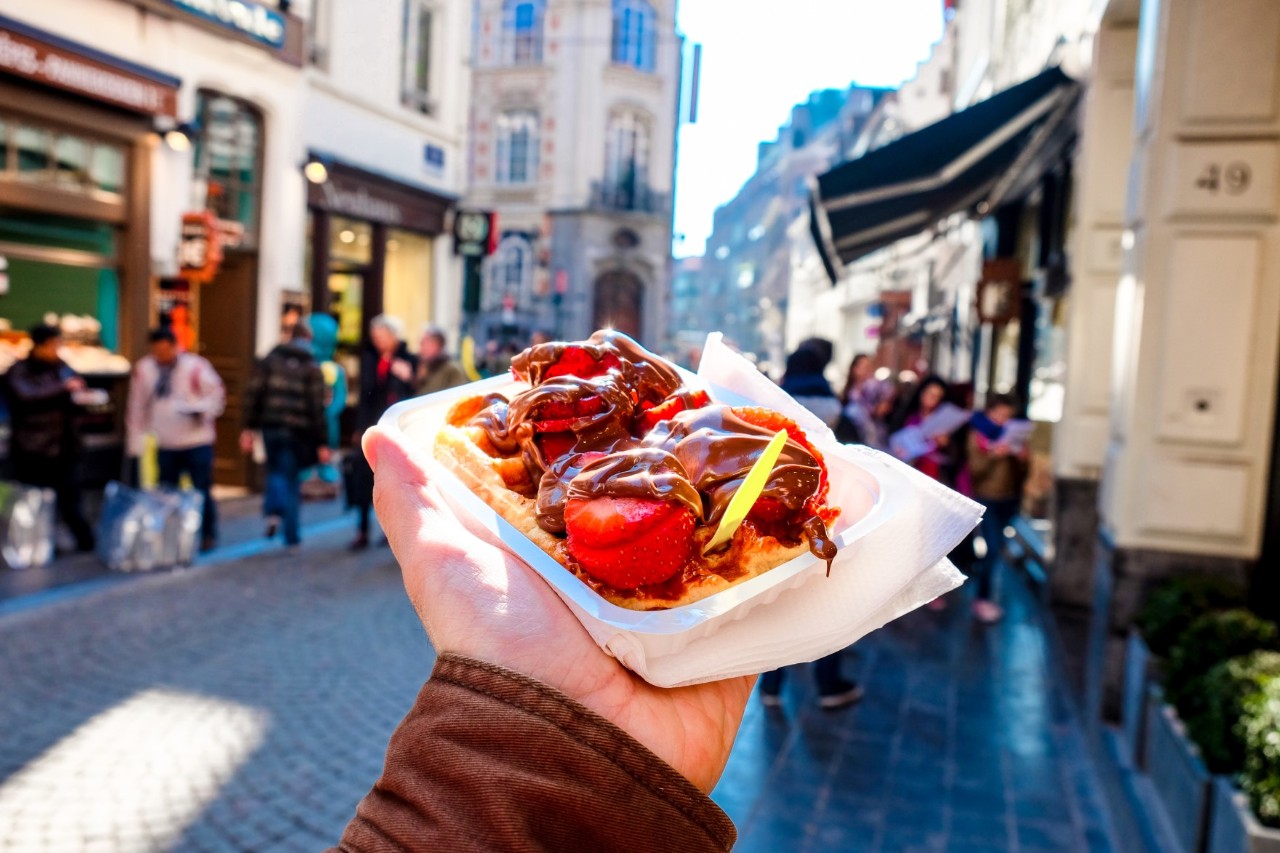 Hand holding Belgian waffles with chocolate, in the background crowd of people, pedestrian zone and shops 