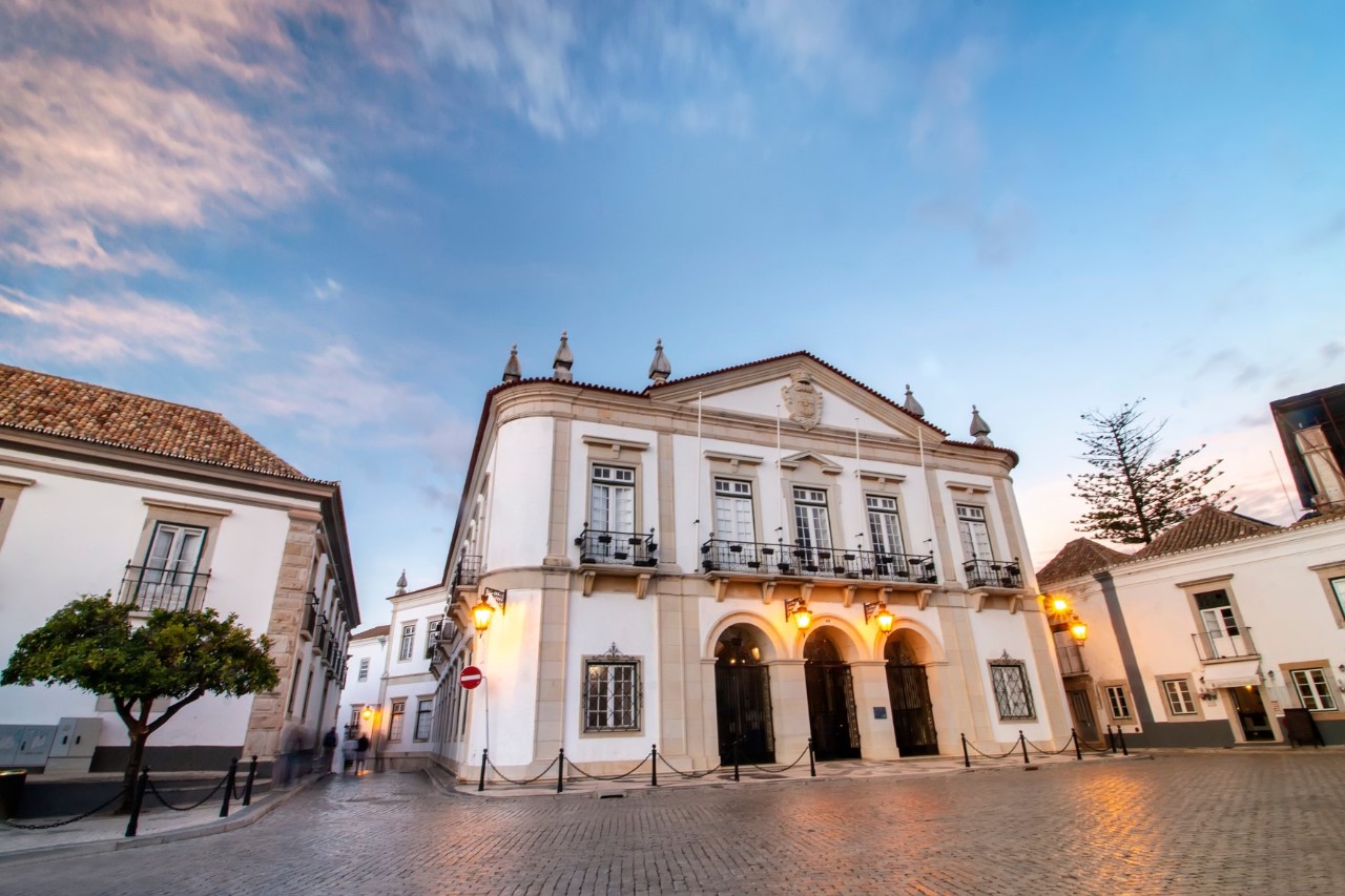 Faro Town Hall in the old town at dusk, empty alley and quiet square surrounded by white houses, yellow lights above the entrance. 