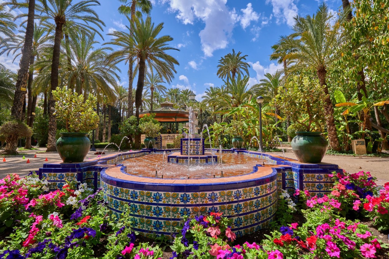 A fountain decorated with blue and colourful mosaic tiles and surrounded by colourful flowers at the bottom. Different types of palm trees can be seen in the background.