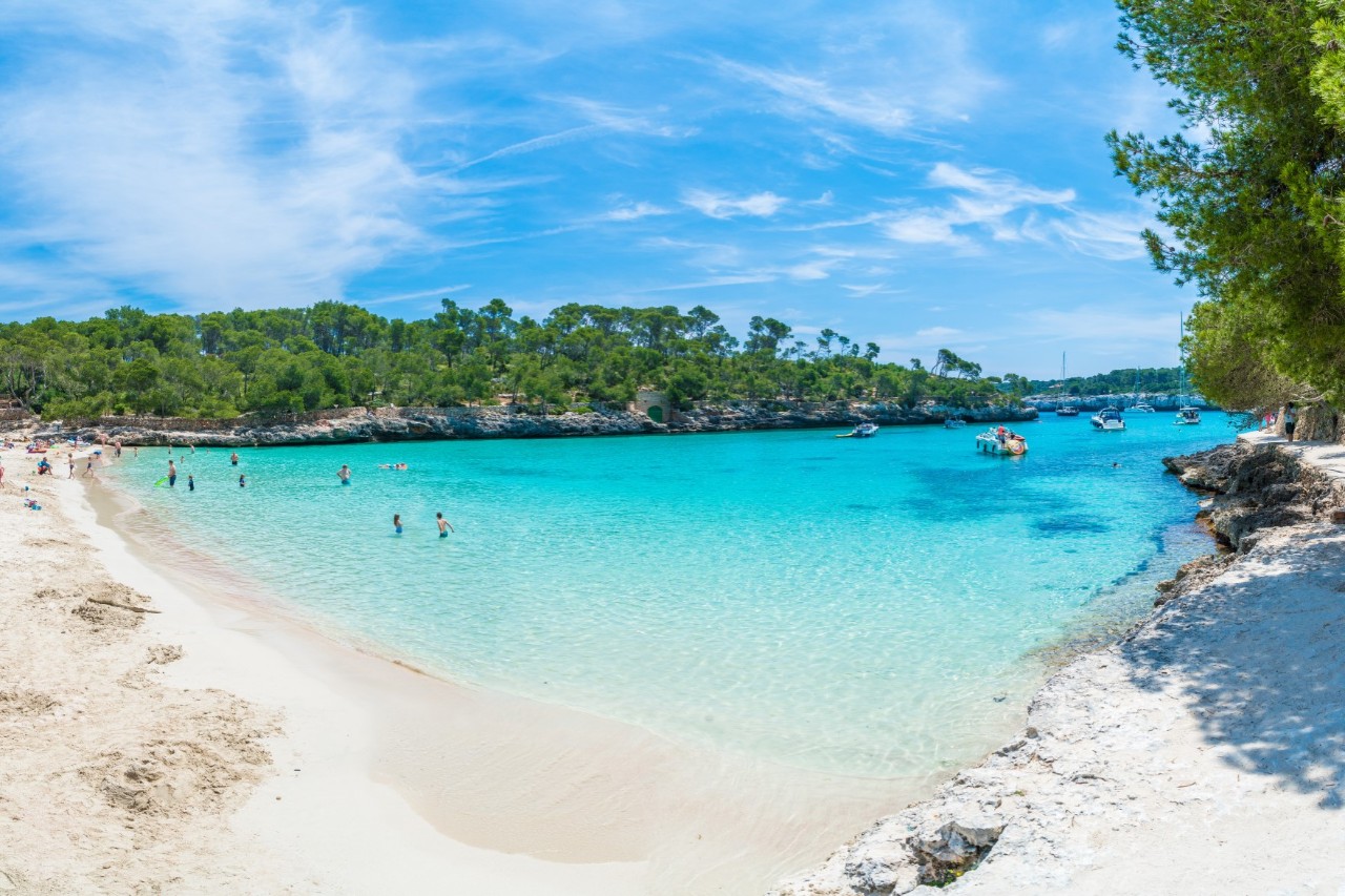 Turquoise bay with a white sandy beach, few bathers and small boats, surrounded by trees 