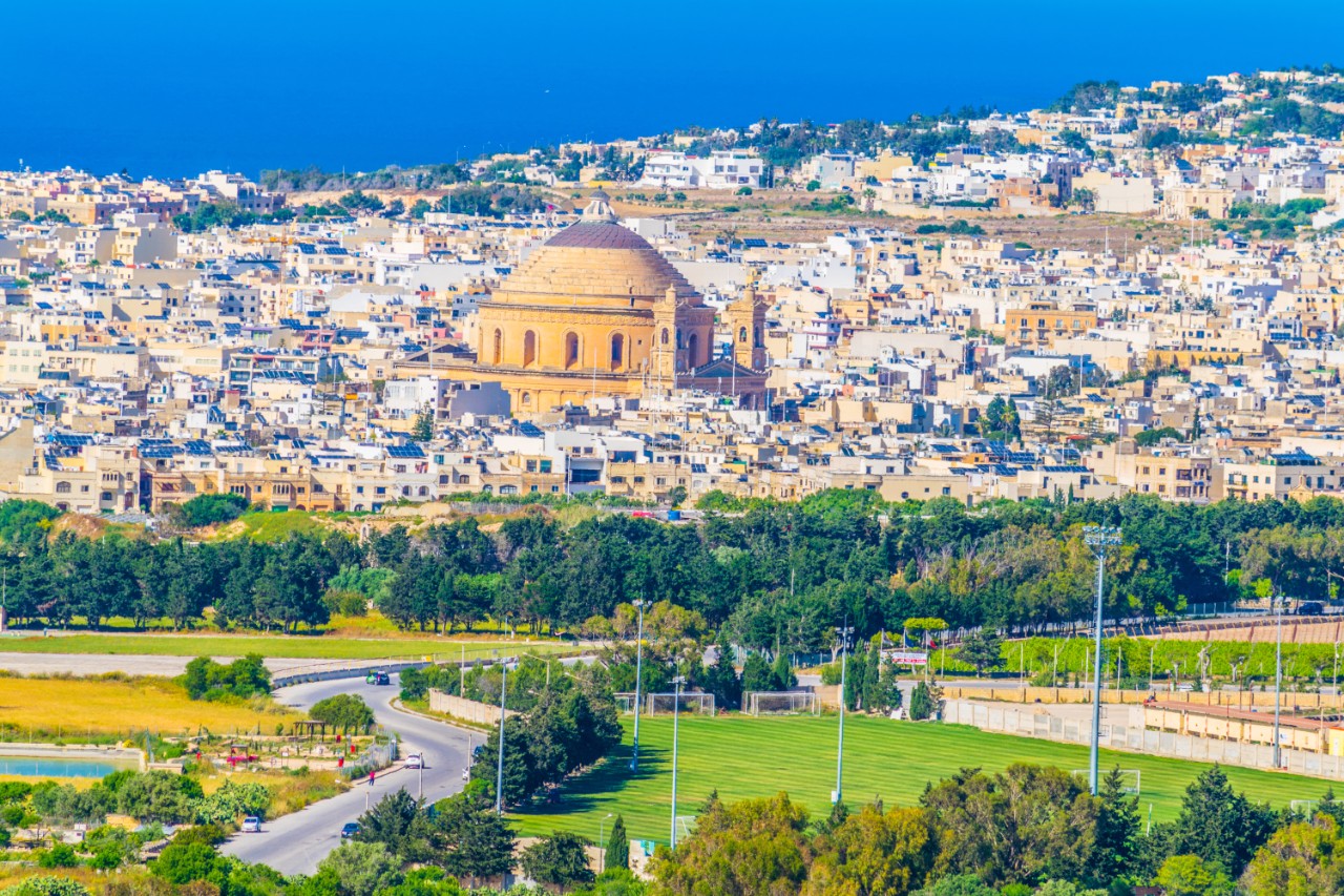 Mosta Rotunda with dome, surrounded by houses, sea in the background.