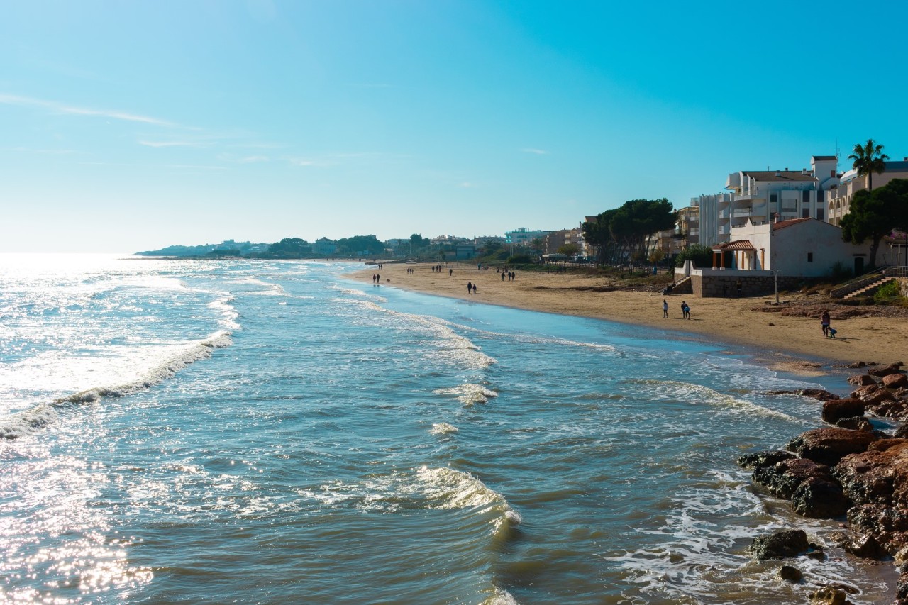 Sandy beach with few people and white buildings by the gently wavy sea 