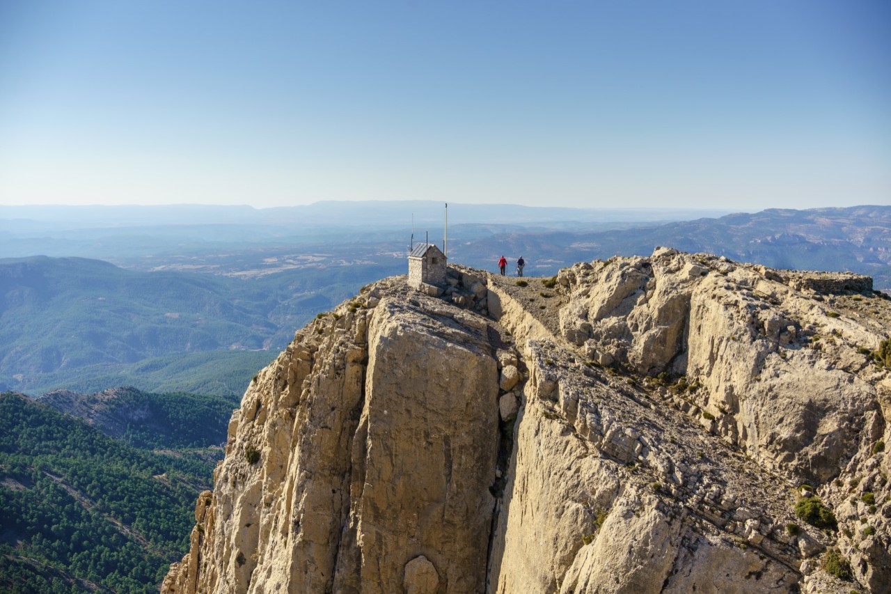 A rocky peak with a steeply sloping face on which two people are standing next to a small hut with a green, extensive valley behind them 