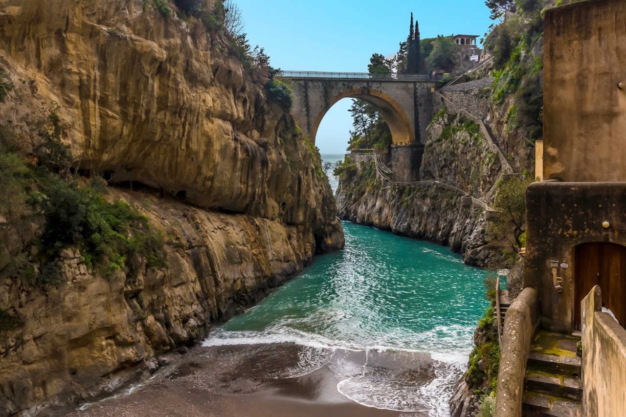 A very narrow bay that leads onto a sandy beach. Steep rocks rise up on the left, a building and a narrow staircase can be seen on the right.