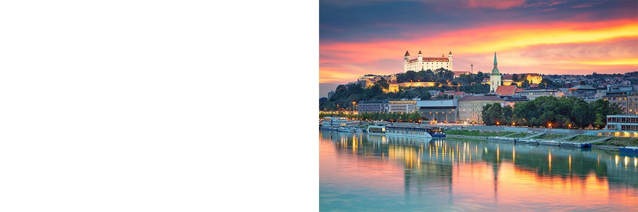 Panoramic view of Bratislava at sunset with the illuminated castle on a hill above the city. In the foreground, the city with its church, waterfront promenade, and ships is reflected in the calm waters of the Danube.