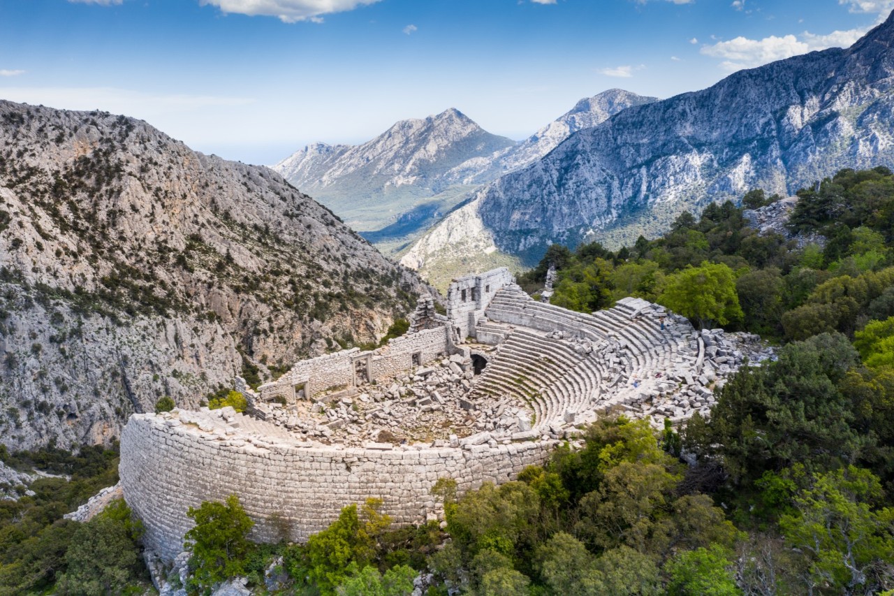 Ancient amphitheatre in the middle of a mountainous landscape. The ruin, made of stone blocks, is surrounded by green trees.