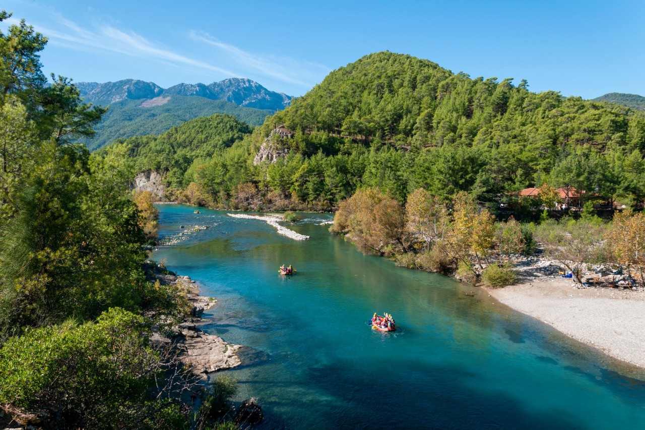River flowing through a green landscape. There are two boats with people in the water. Forested hills and mountains rise in the background.