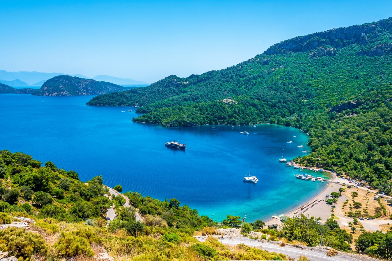 The picture shows a bay with turquoise blue water surrounded by densely wooded hills. Two boats are anchored in the bay.