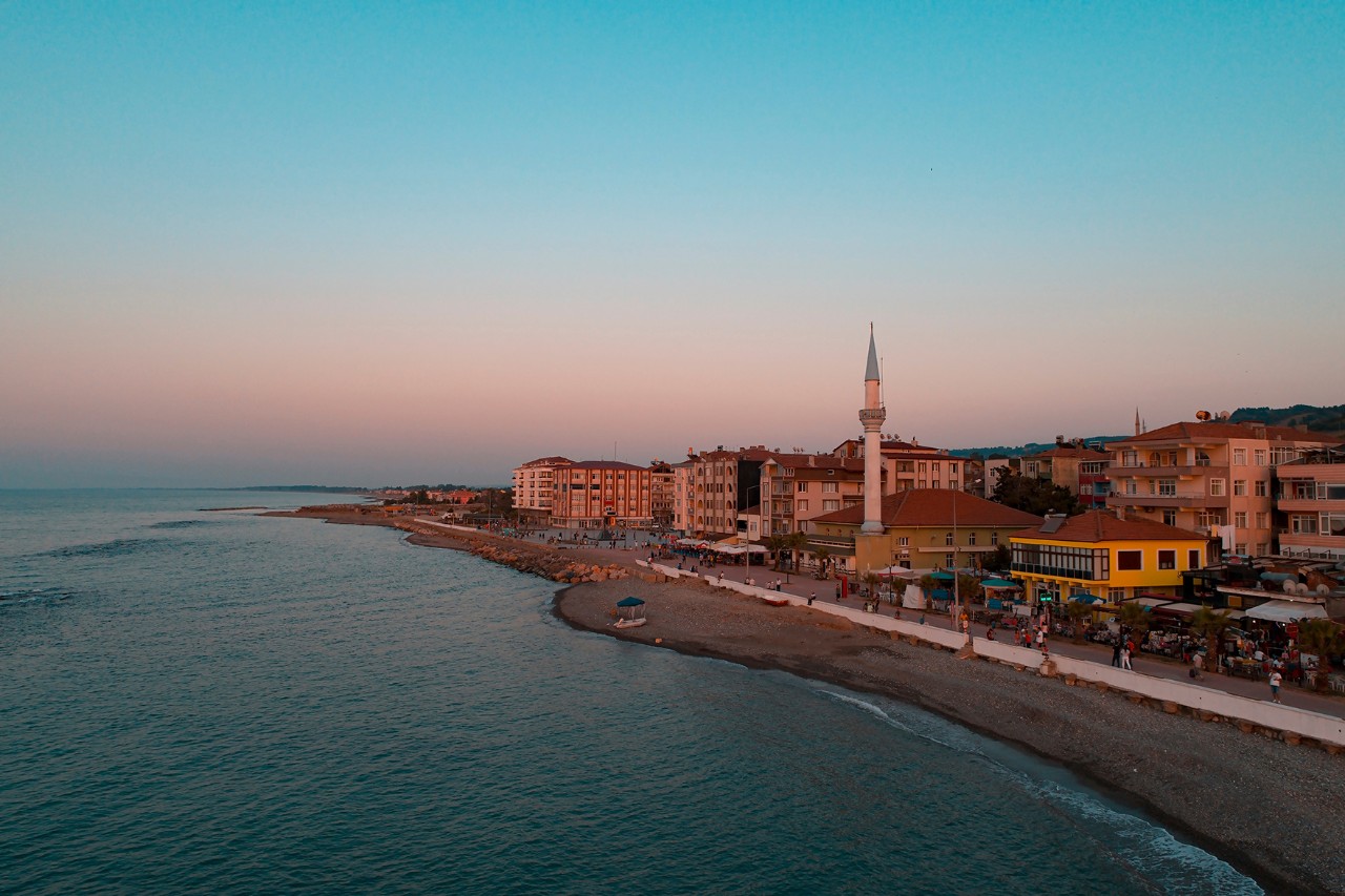 Coastal promenade at sunset with a mosque and a tall minaret in the center. Several houses line the beach. 