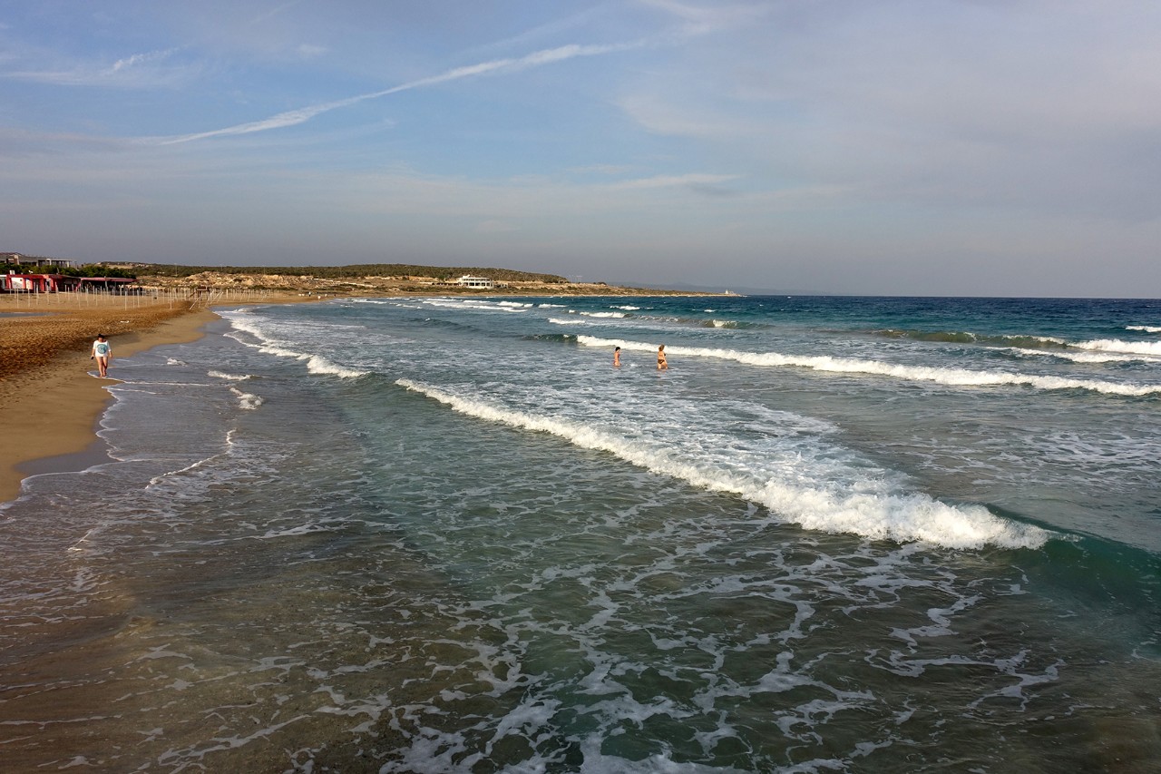 Empty, long sandy beach with gentle waves and calm sea under a slightly overcast sky.
