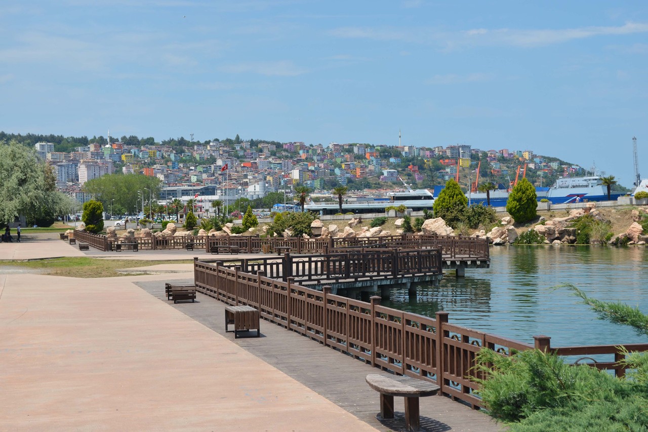 Waterfront promenade with wooden walkways and benches on a tranquil body of water. In the background, a colorful, densely built-up city can be seen on a hill, as well as a harbor with several ships.