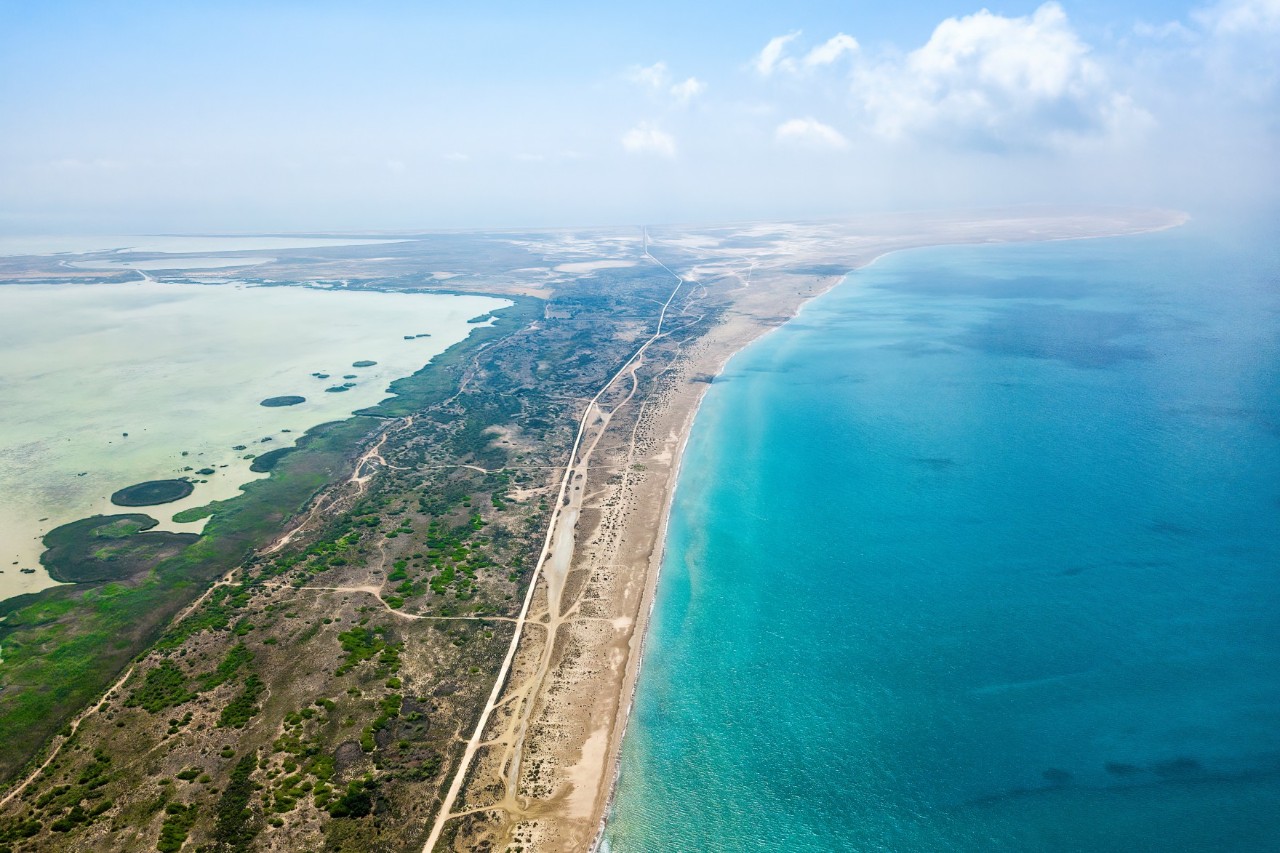 Aerial view of a coastal landscape with turquoise blue sea on the right and shallow lagoons on the left. A sandy strip, partly covered with green vegetation, separates the two bodies of water.