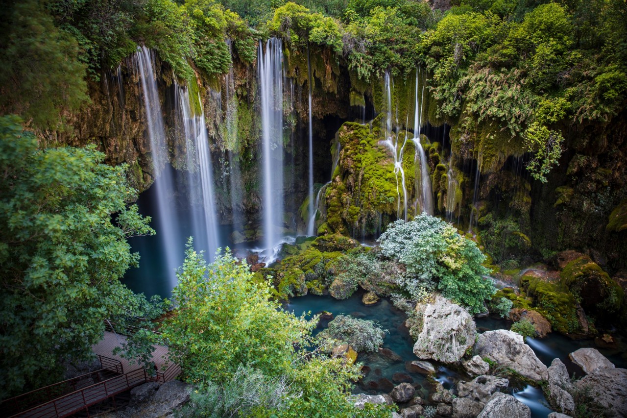 Several narrow waterfalls plunge into a turquoise blue pool surrounded by large rocks and dense green vegetation.