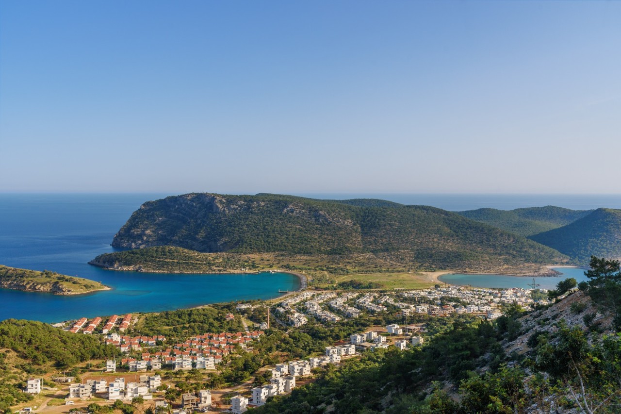 Panoramic view of a picturesque coastal landscape with a turquoise blue bay surrounded by green hills. In the foreground, white houses can be seen nestled among green vegetation.