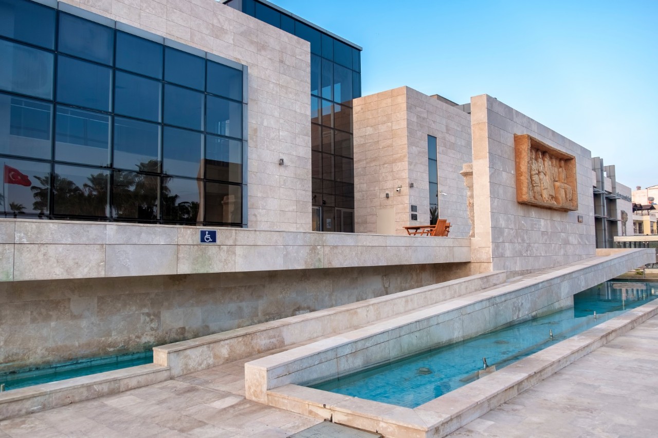 Modern building with a light-colored natural stone façade and large glass windows. The façade features a bas-relief depicting historical figures. A shallow water channel runs in front of the building.