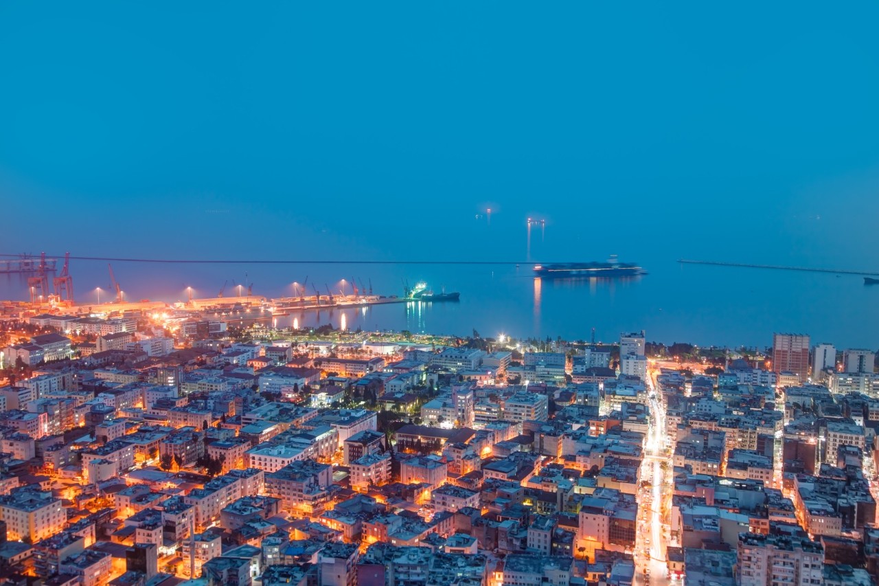 City view with illuminated buildings, harbor facilities, and ships on the calm sea under a deep blue sky in the evening light.