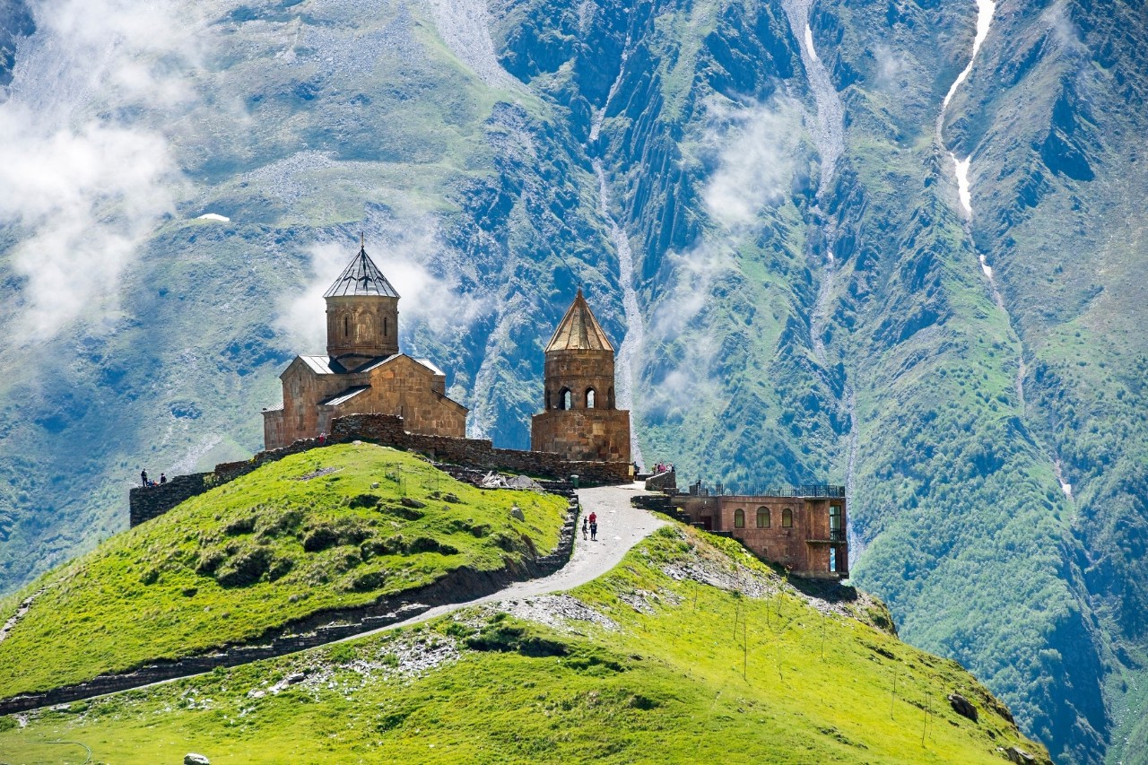 A church standing on a green hill. A narrow path runs in front of it. In the background, huge wooded mountain slopes with veils of clouds can be seen.