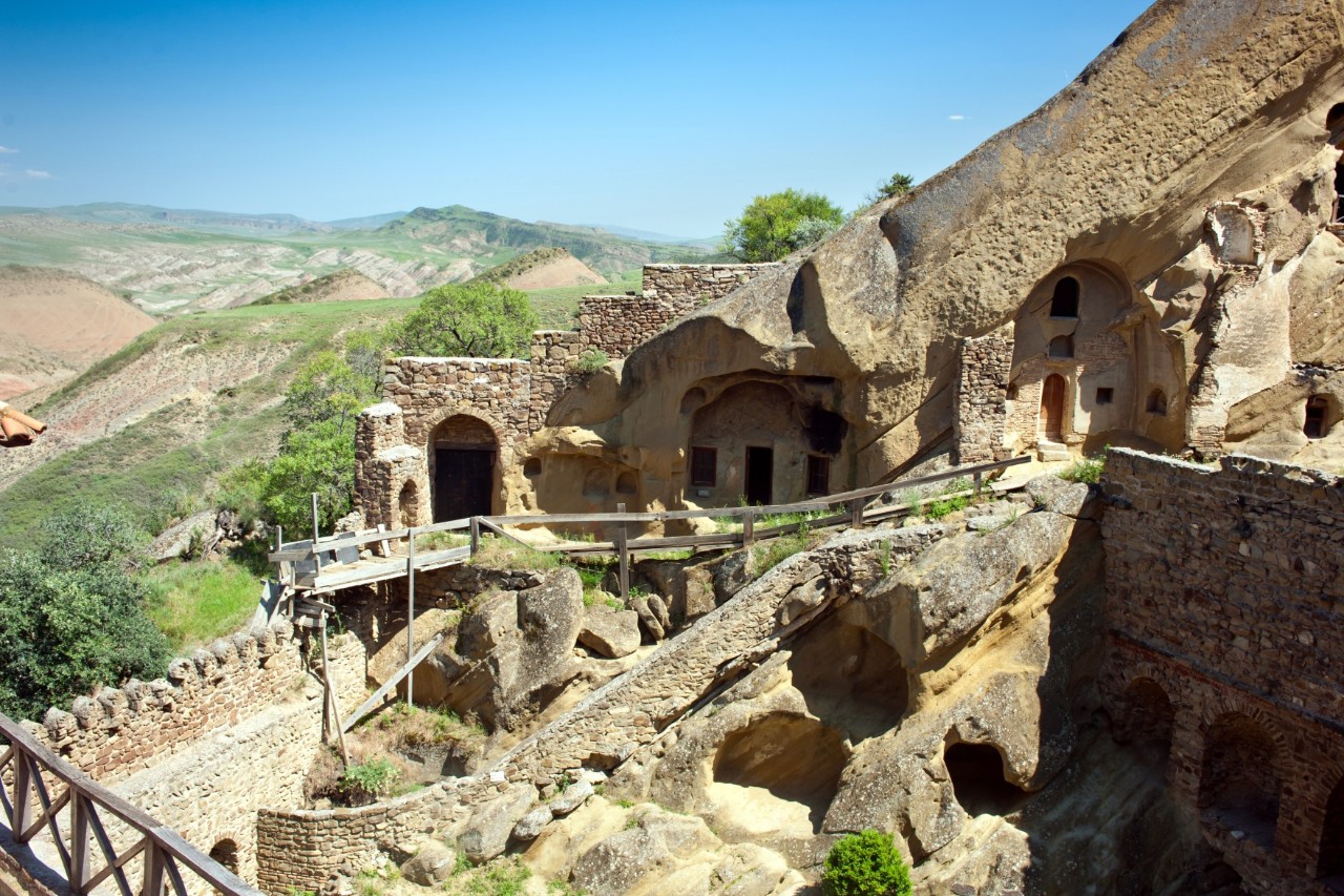 A monastery complex built into the rocks, with doors and windows, stands before a vast landscape stretching out under a clear blue sky.