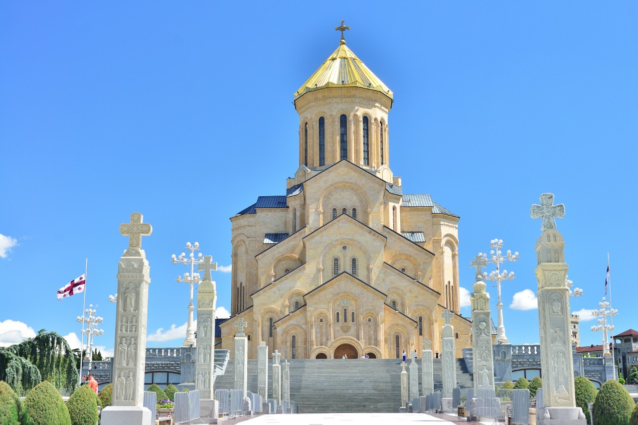 From a low angle, a cathedral with a golden dome and monumental façade can be seen standing on a spacious square. Columns topped with crosses lead towards the building. Trees frame the image on the left and right. The sky is bright blue.