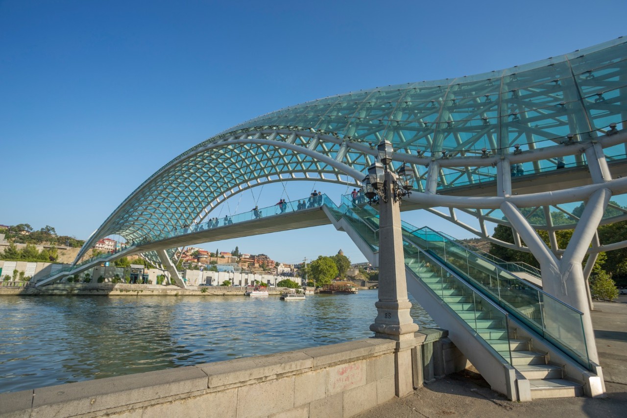 View of a glass, wave-shaped pedestrian bridge spanning diagonally across a body of water. Several people can be seen on the bridge. In the background, a city lies on the shore under a clear blue sky.