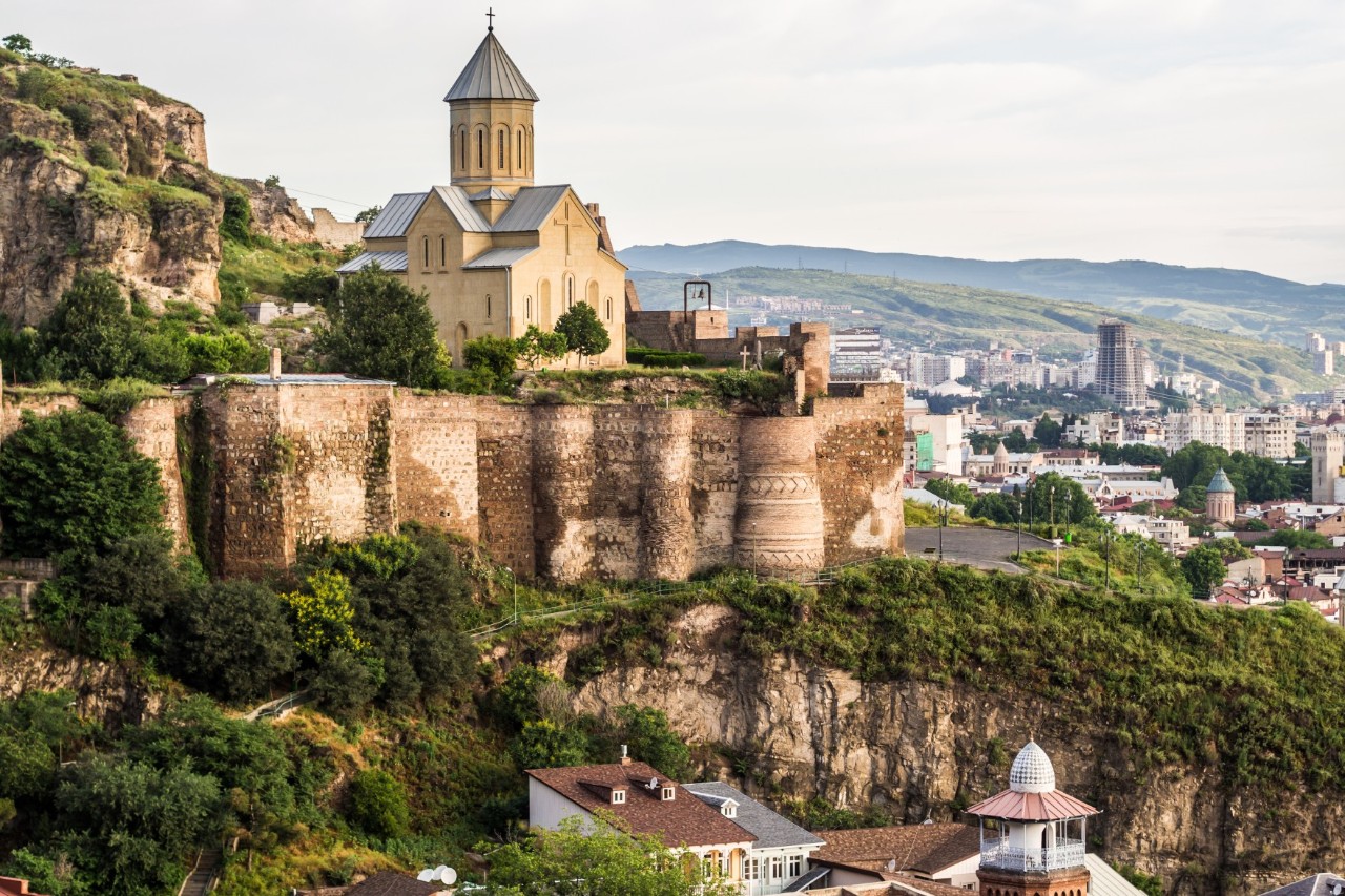 A fortress sits on a green rock above the town. In front of it is a church. In the foreground are rooftops, and in the background, the town spreads out into the hilly landscape.