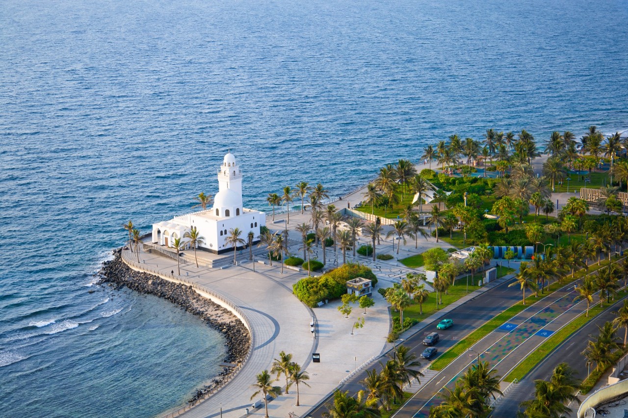 A white mosque directly on the coast, water on the left, palm trees and a six-lane road on the right. 
